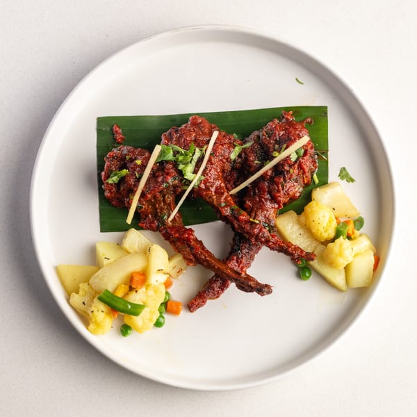 Red chicken lollipops on a banana leaf, garnished with ginger and cilantro, served with mixed vegetables.