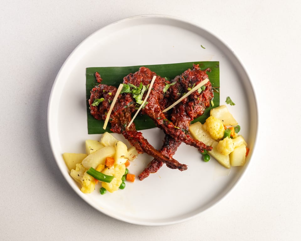 Red chicken lollipops on a banana leaf, garnished with ginger and cilantro, served with mixed vegetables.