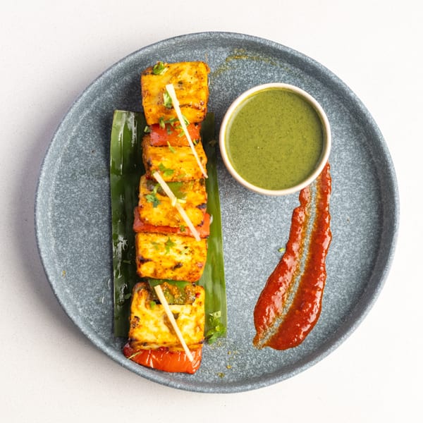 Grilled paneer and vegetable skewers with green chutney and red sauce on a gray plate.