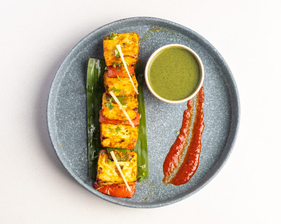 Grilled paneer and vegetable skewers with green chutney and red sauce on a gray plate.
