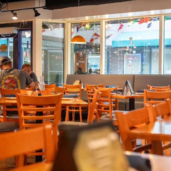 Restaurant interior with wooden tables and chairs, a couple dining, and large windows overlooking a concourse.