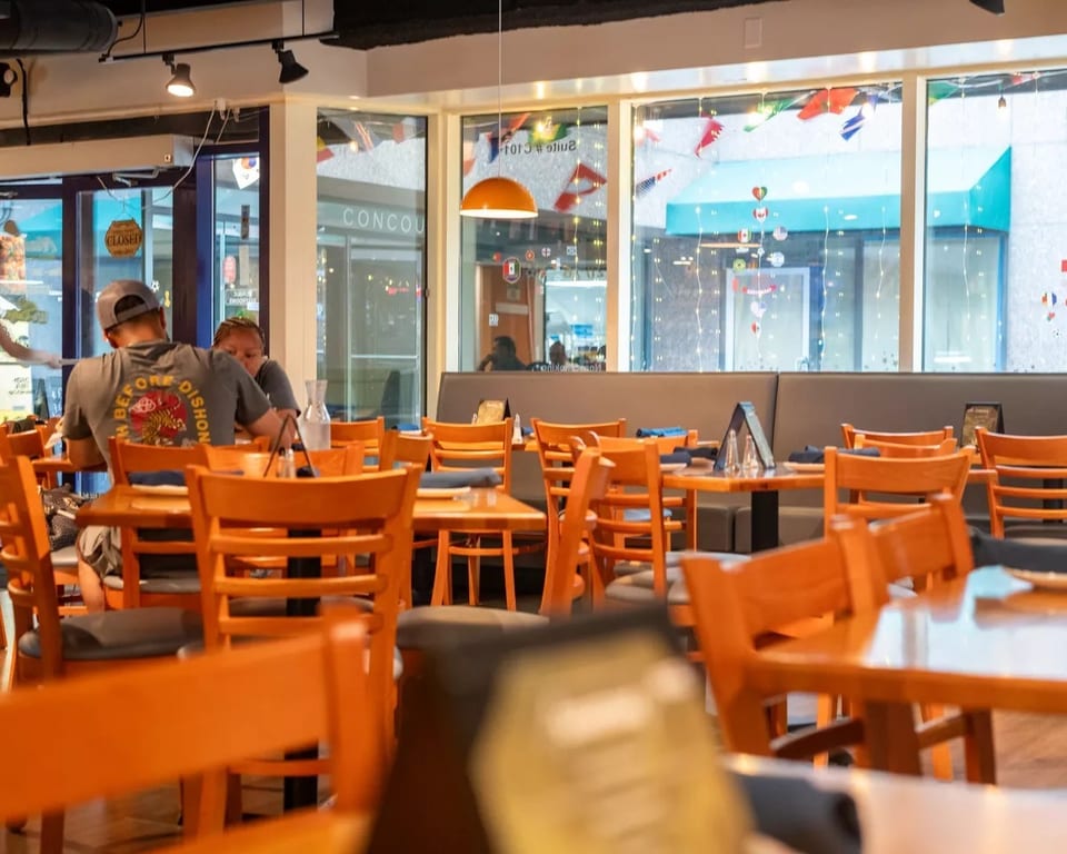 Restaurant interior with wooden tables and chairs, a couple dining, and large windows overlooking a concourse.