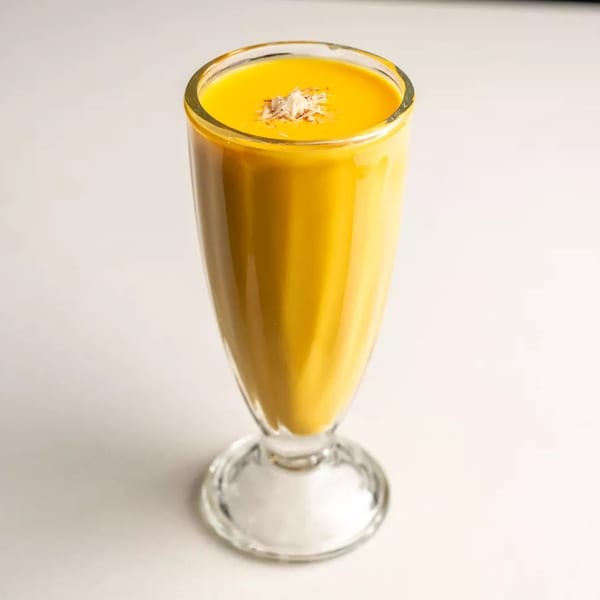 A tall glass of yellow mango lassi, topped with shredded coconut, on a white surface.