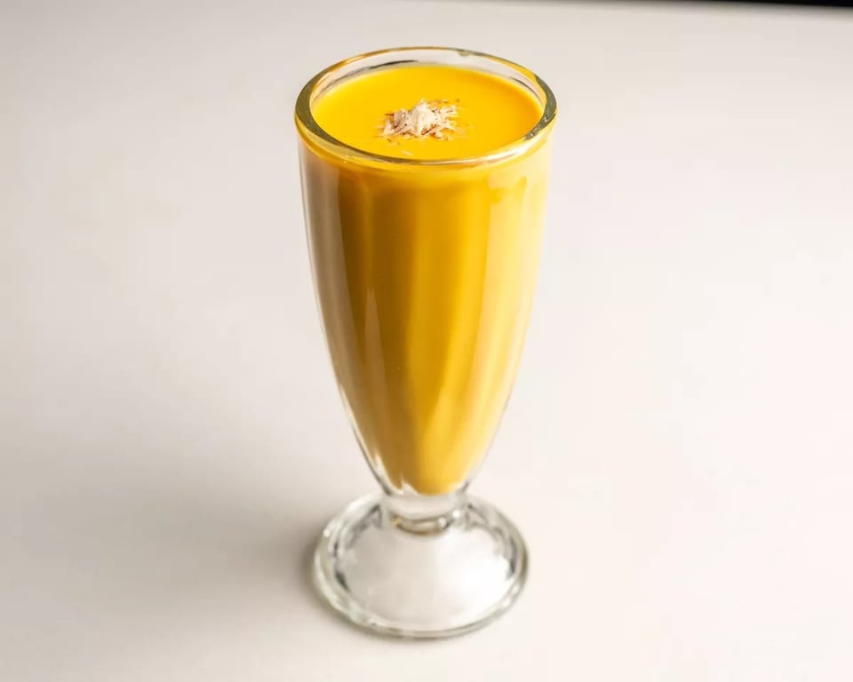 A tall glass of yellow mango lassi, topped with shredded coconut, on a white surface.