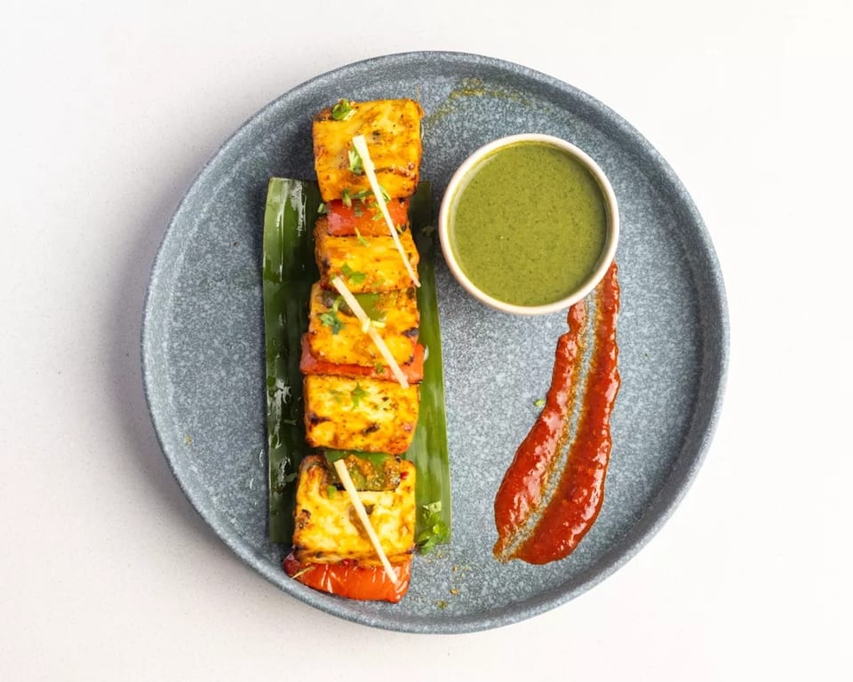 Grilled paneer and vegetable skewers with green chutney and red sauce on a grey plate.