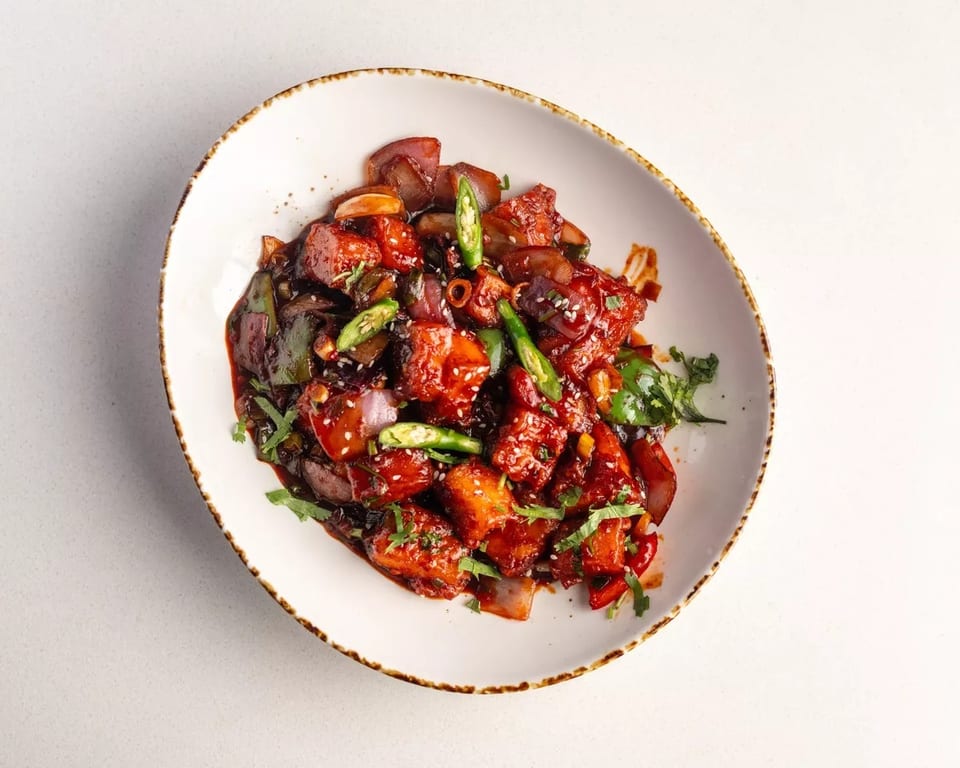 Chili Paneer with red sauce, green chilies, onions, and cilantro in a white bowl.