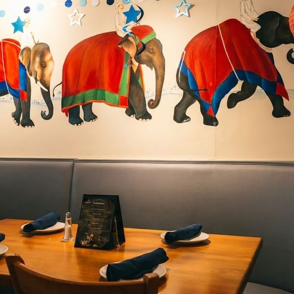 Restaurant interior with elephant mural, blue booth seating, and wooden tables set with plates and napkins.