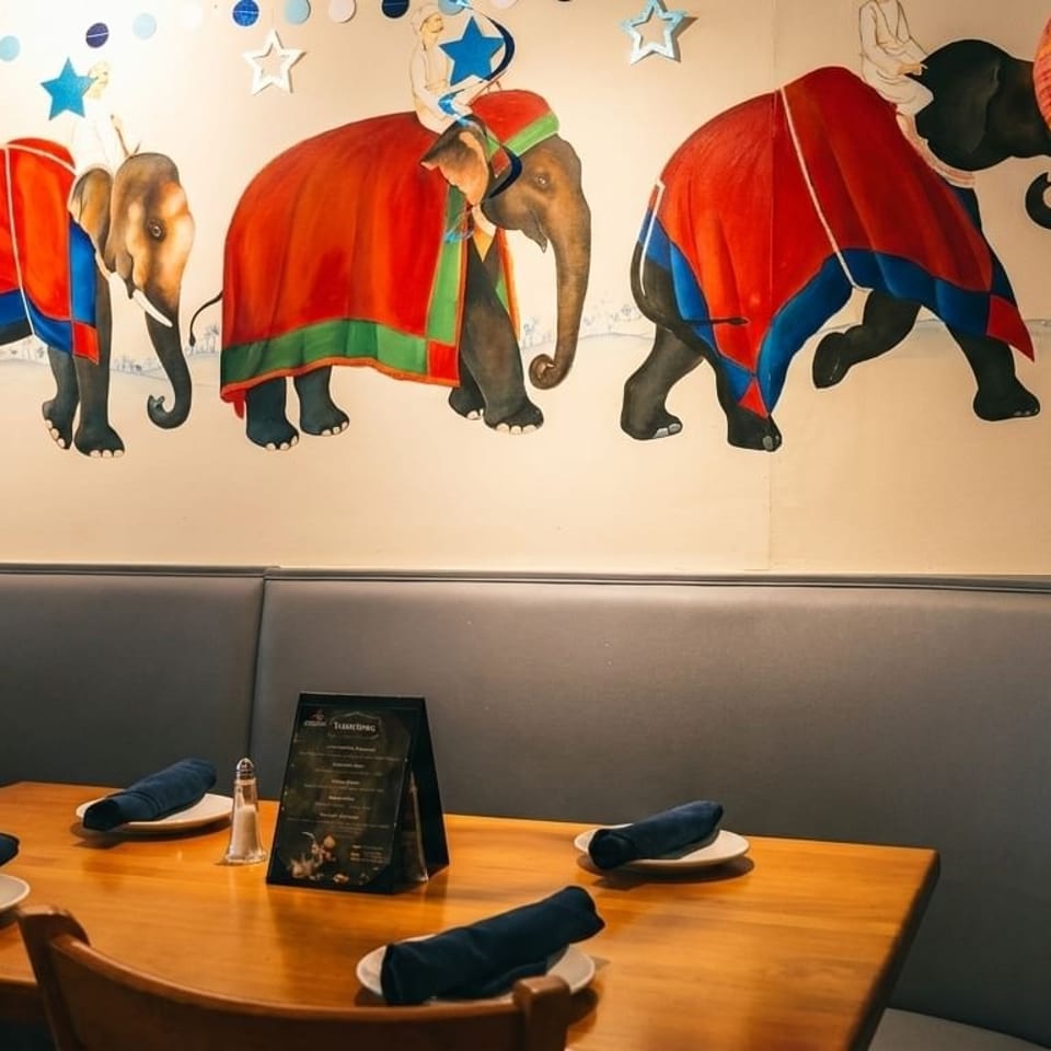 Restaurant interior with elephant mural, blue booth seating, and wooden tables set with plates and napkins.