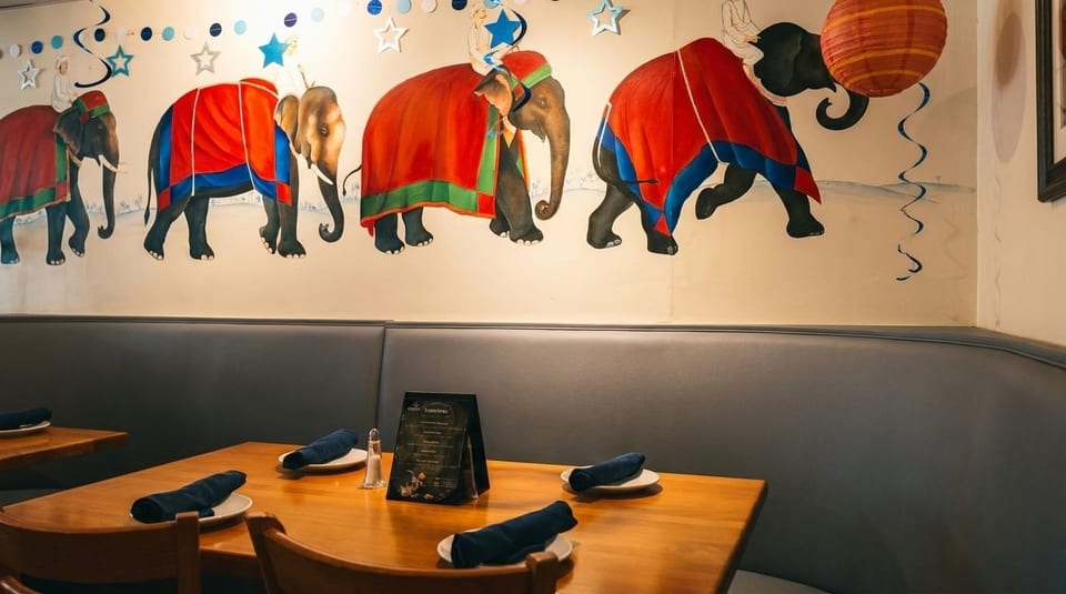 Restaurant interior with elephant mural, blue booth seating, and wooden tables set with plates and napkins.