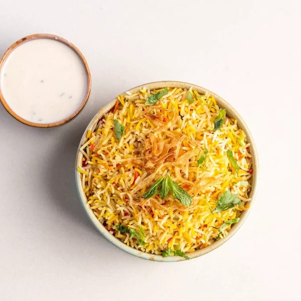 Overhead view of a bowl of biryani rice with fried onions and mint, next to a small bowl of raita.