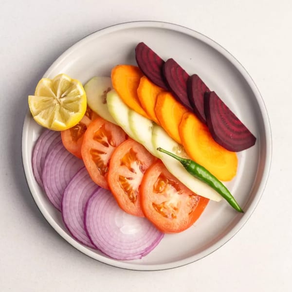A plate of sliced red onions, tomatoes, cucumber, carrots, beets, a lemon wedge, and a green chili.