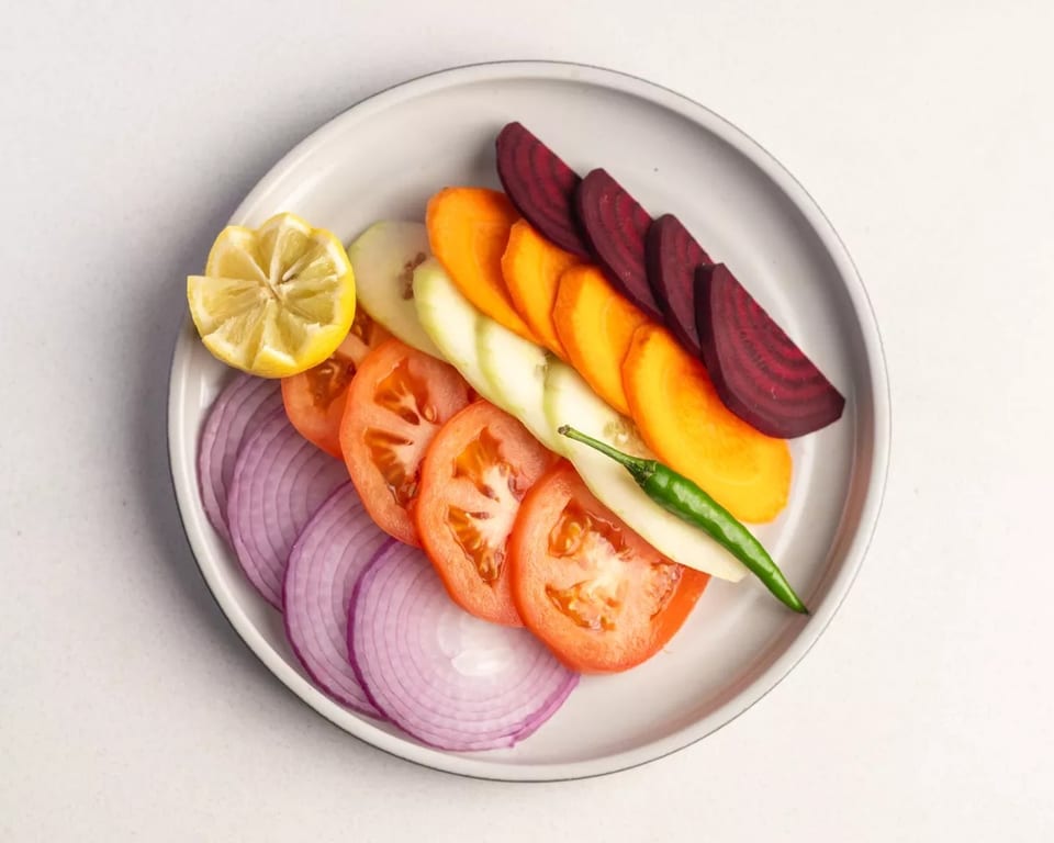 A plate of sliced red onions, tomatoes, cucumber, carrots, beets, a lemon wedge, and a green chili.