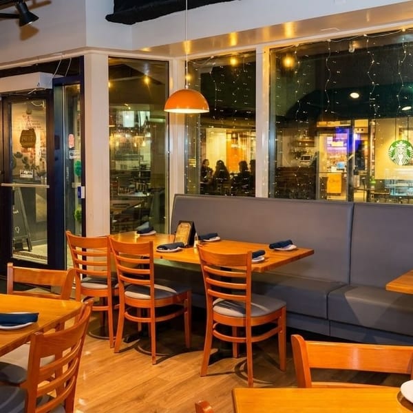 Empty restaurant dining room with wooden tables, chairs, and a long booth next to large windows.